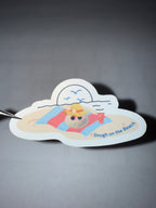 Sticker with a beach scene and a ball of bread dough on a beach towel wearing sunglasses and a sunhat, featuring the pun "dough on the beach"
