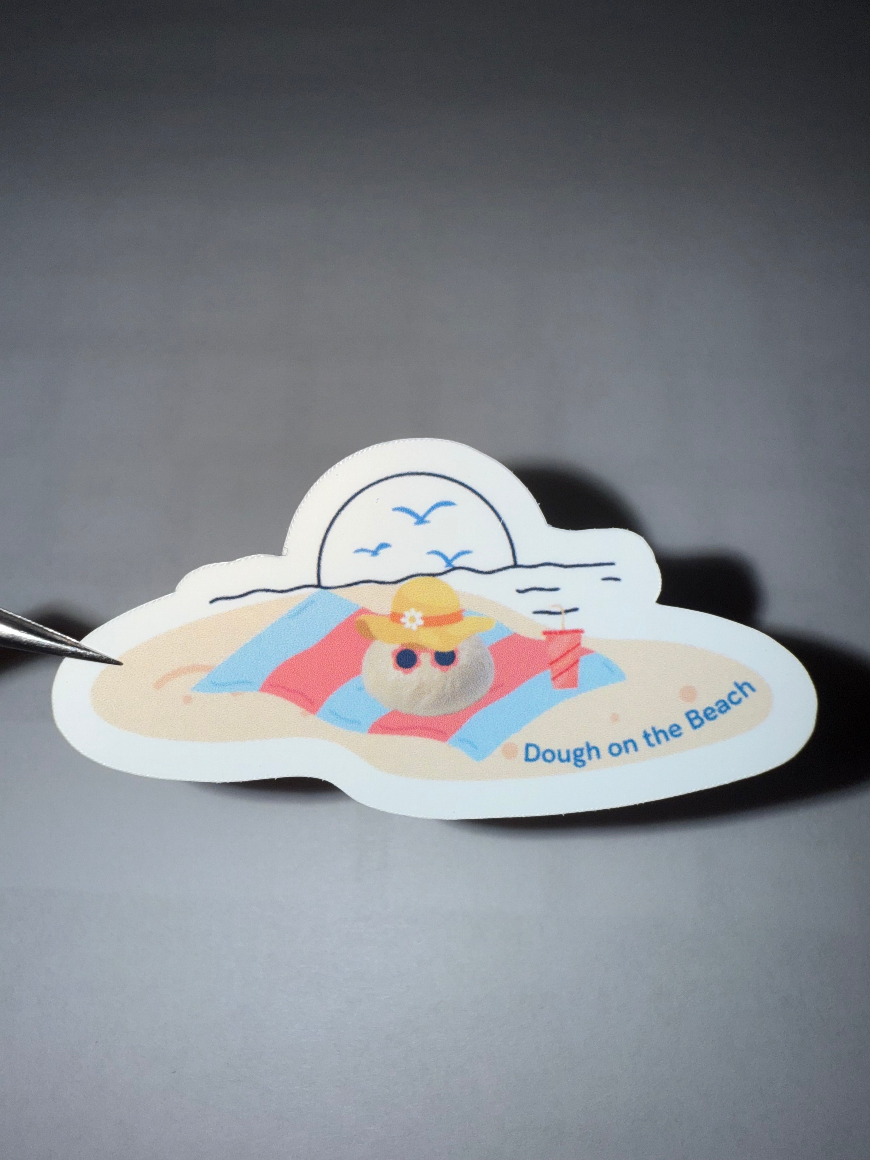 Sticker with a beach scene and a ball of bread dough on a beach towel wearing sunglasses and a sunhat, featuring the pun "dough on the beach"