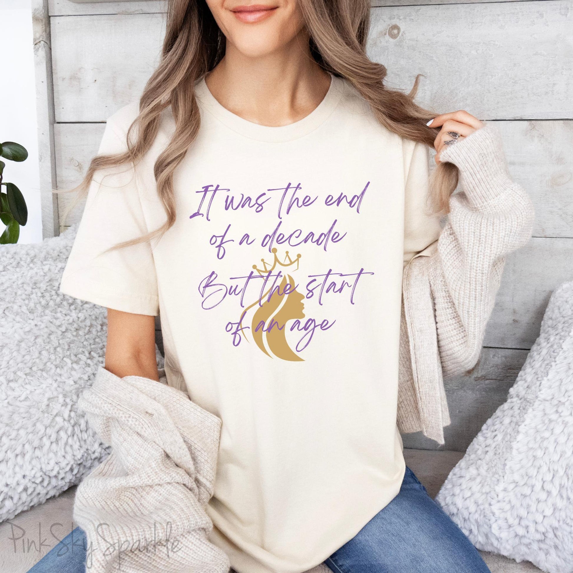 Embrace the excitement of a new era with our End of a Decade, Start of an Age T-shirt, inspired by Taylor Swift's iconic lyrics. Featuring the words It was the end of a decade, But the start of an age in a purple marker script font with a gold silhouette of a woman wearing a crown.