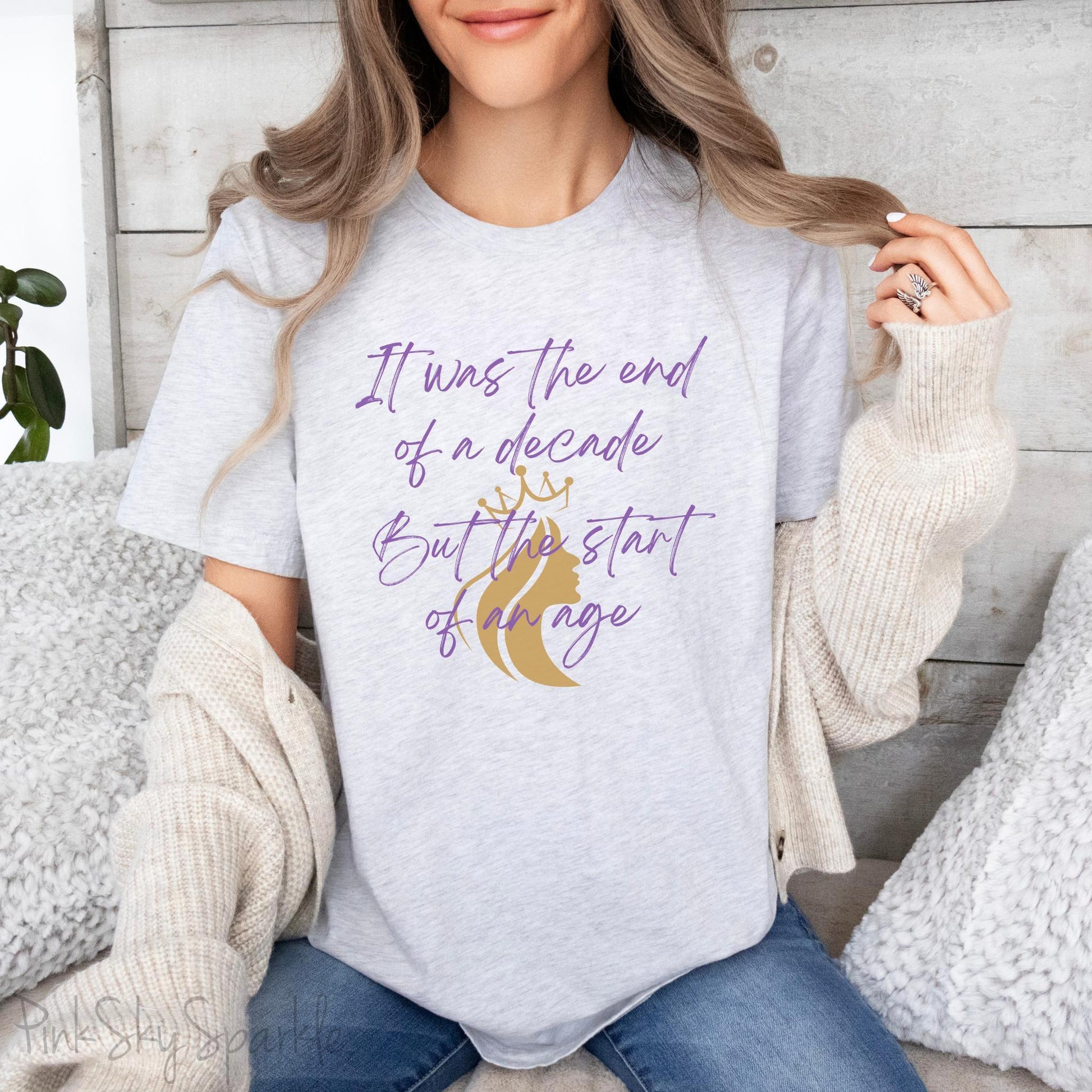 Embrace the excitement of a new era with our End of a Decade, Start of an Age T-shirt, inspired by Taylor Swift's iconic lyrics. Featuring the words It was the end of a decade, But the start of an age in a purple marker script font with a gold silhouette of a woman wearing a crown.