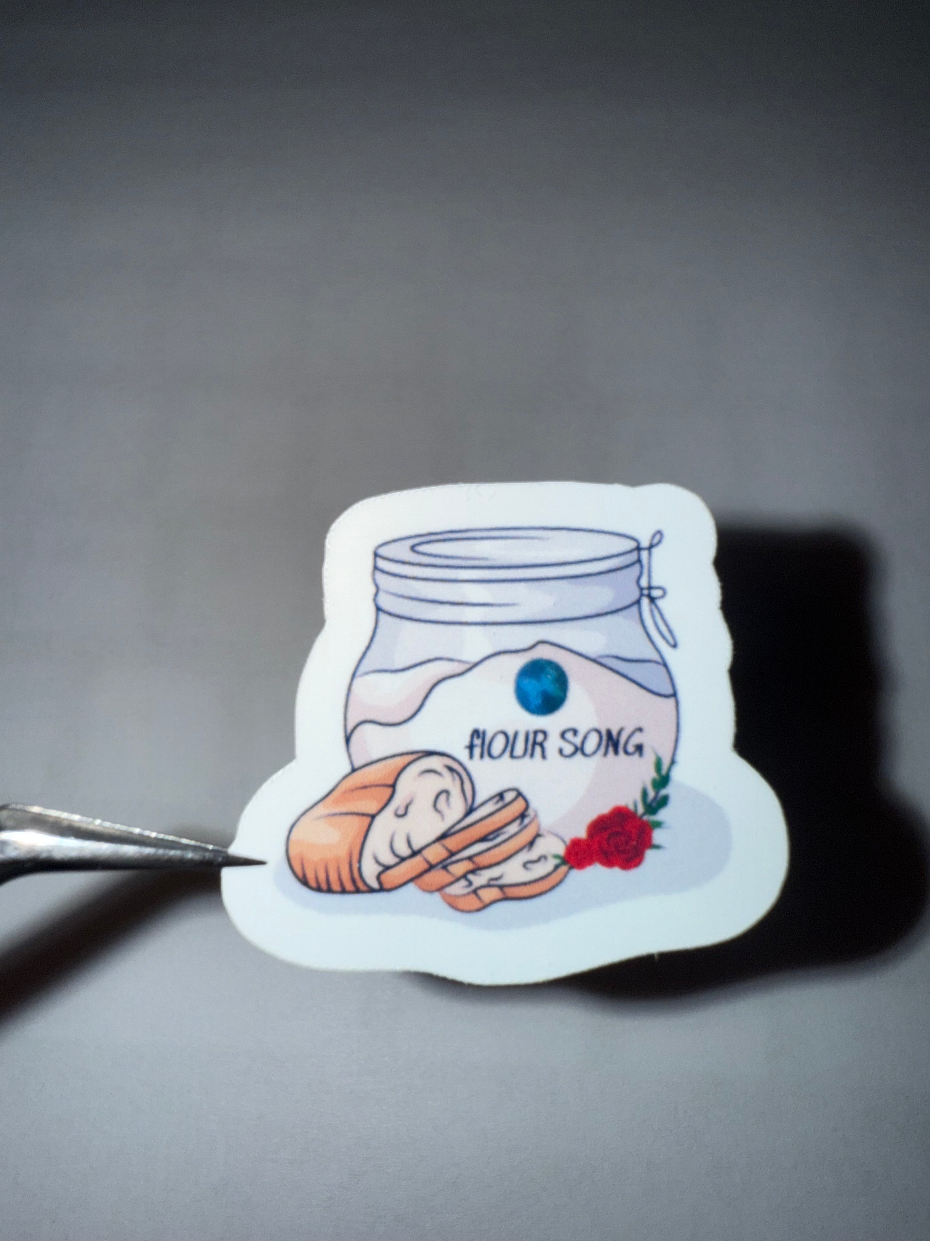 Sticker with jar of flour, loaf of bread and a rose with words "flOUR SONG" on it. 