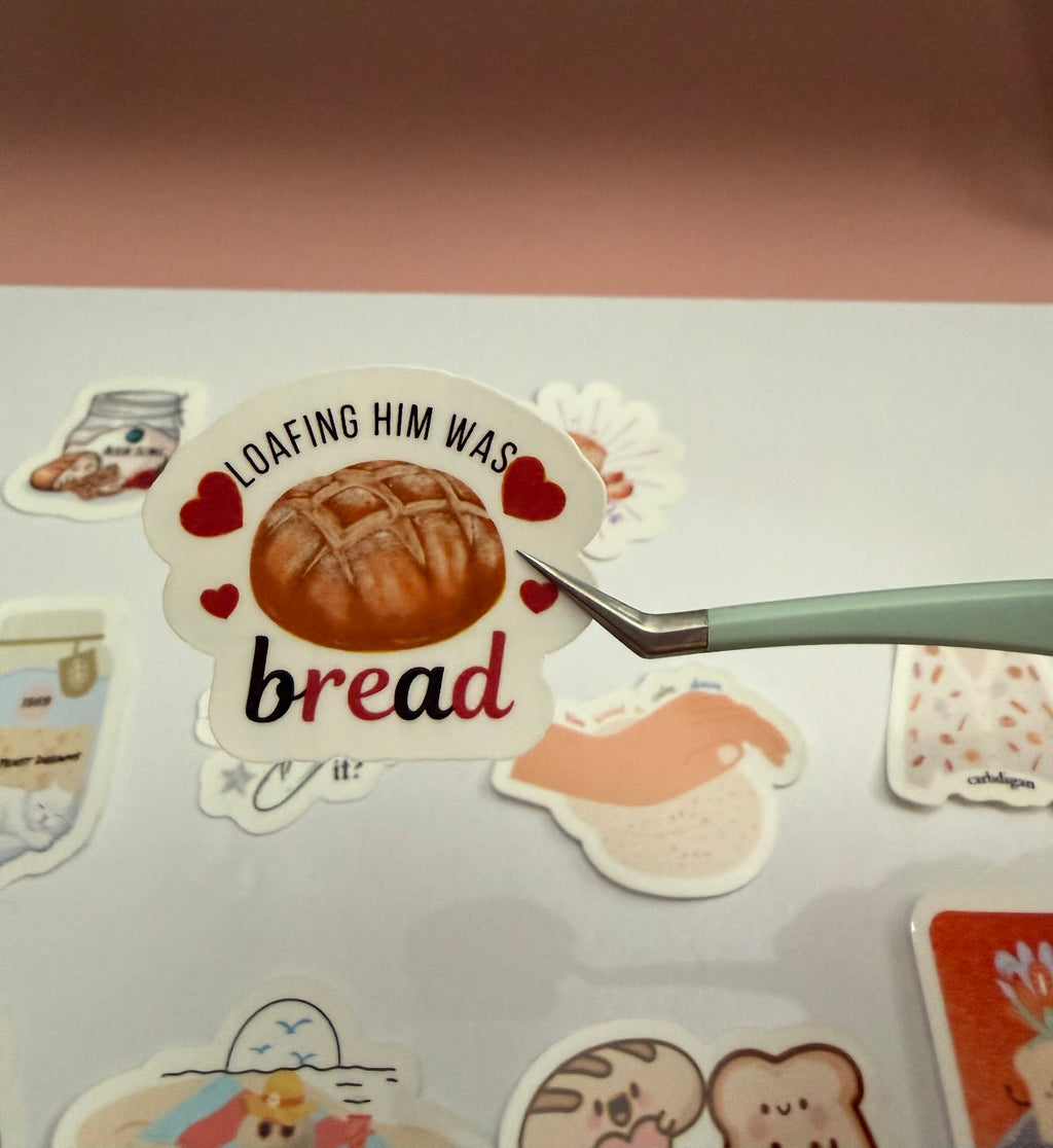 Sticker with bread design and text 'Loafing him was bread' held by a hand on a light surface.