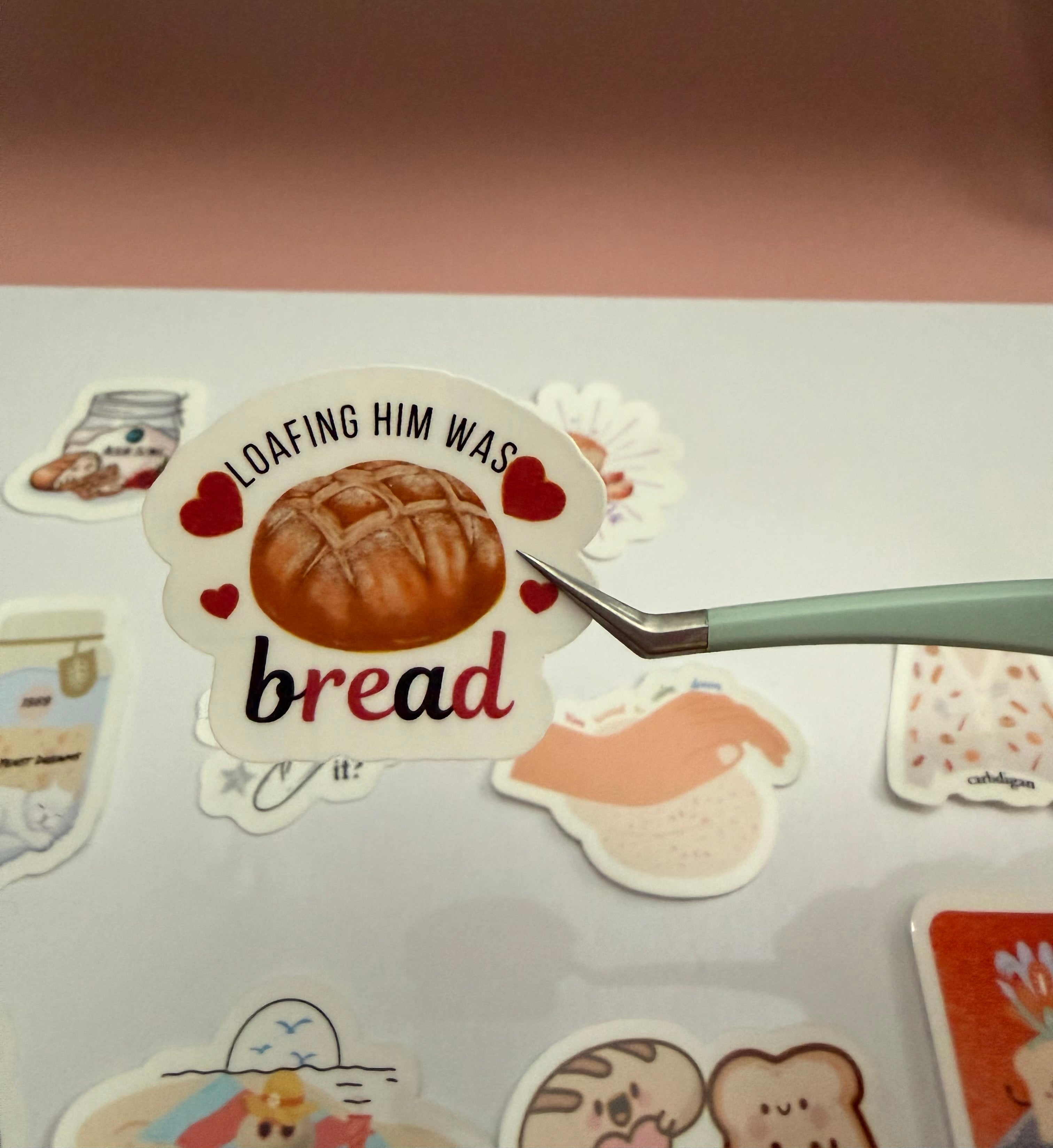 Sticker with bread design and text 'Loafing him was bread' held by a hand on a light surface.