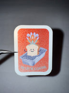 Orange faux-glittery sticker with a cute bread slice wearing a showgirl feather headpiece sitting in a loaf pan  full of diamonds with the pun "the life of a loafgirl"