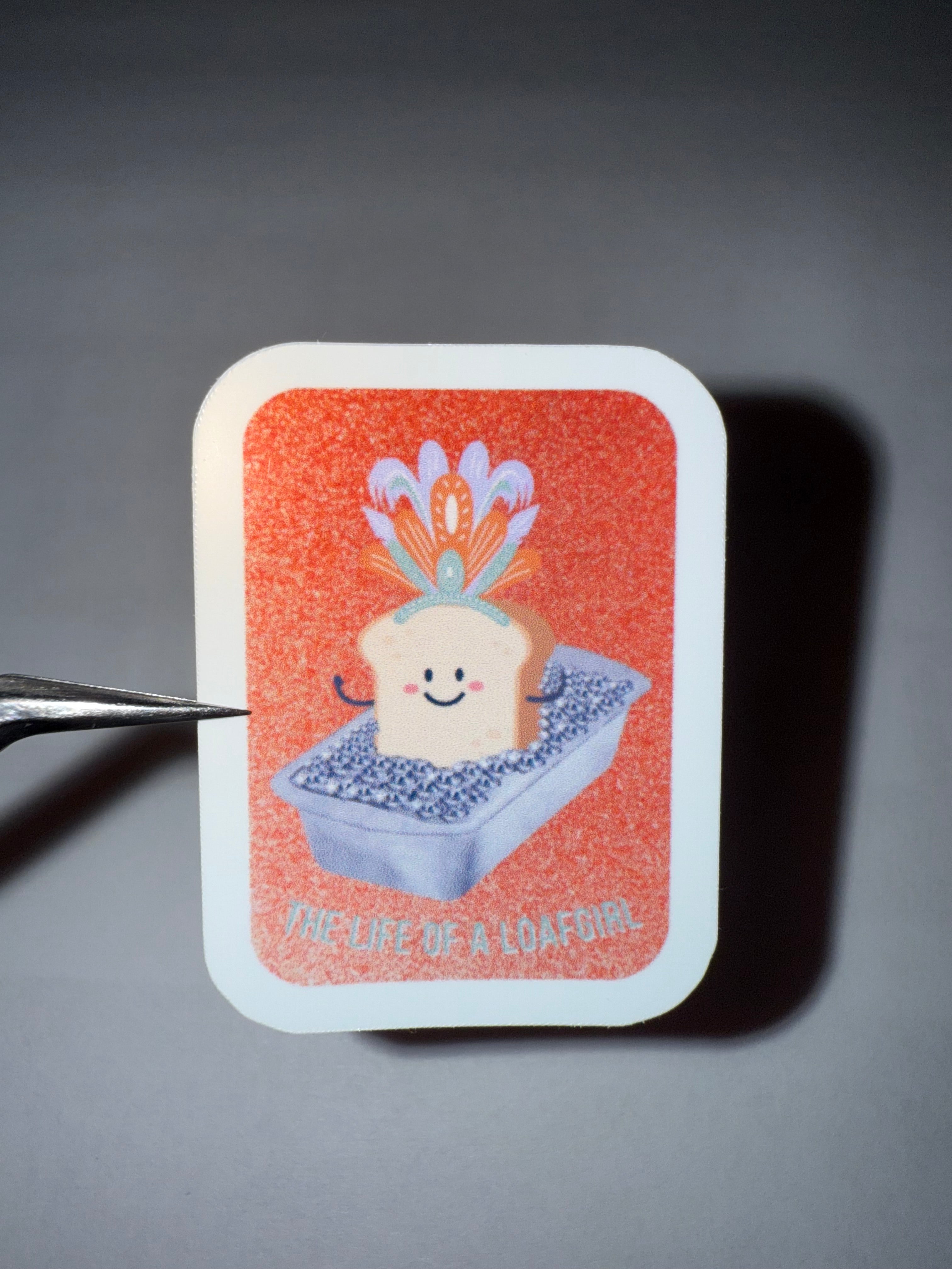 Orange faux-glittery sticker with a cute bread slice wearing a showgirl feather headpiece sitting in a loaf pan  full of diamonds with the pun "the life of a loafgirl"