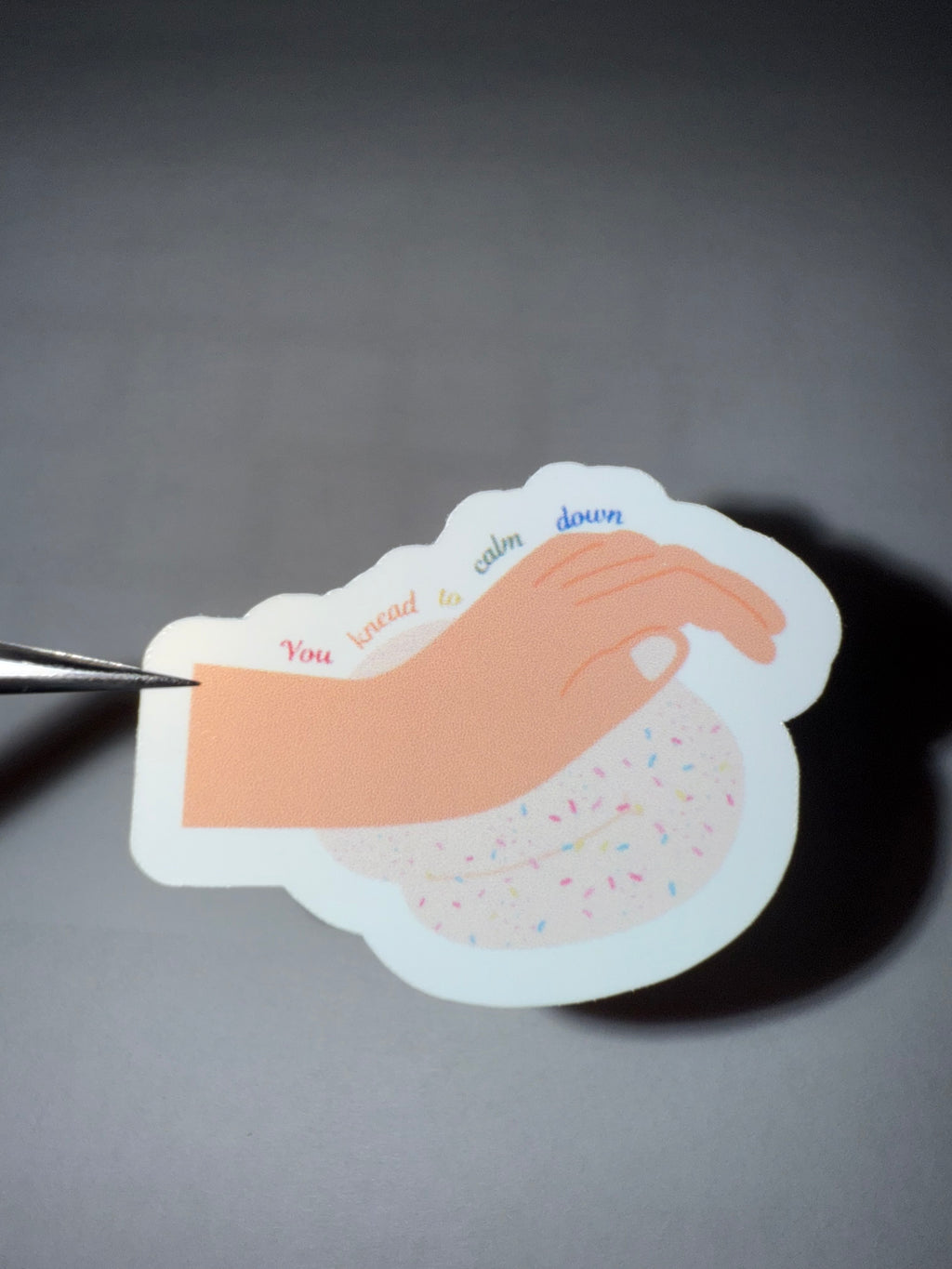 Sticker featuring a hand kneading bread dough with sprinkles with the pun "you knead to calm down" in rainbow font